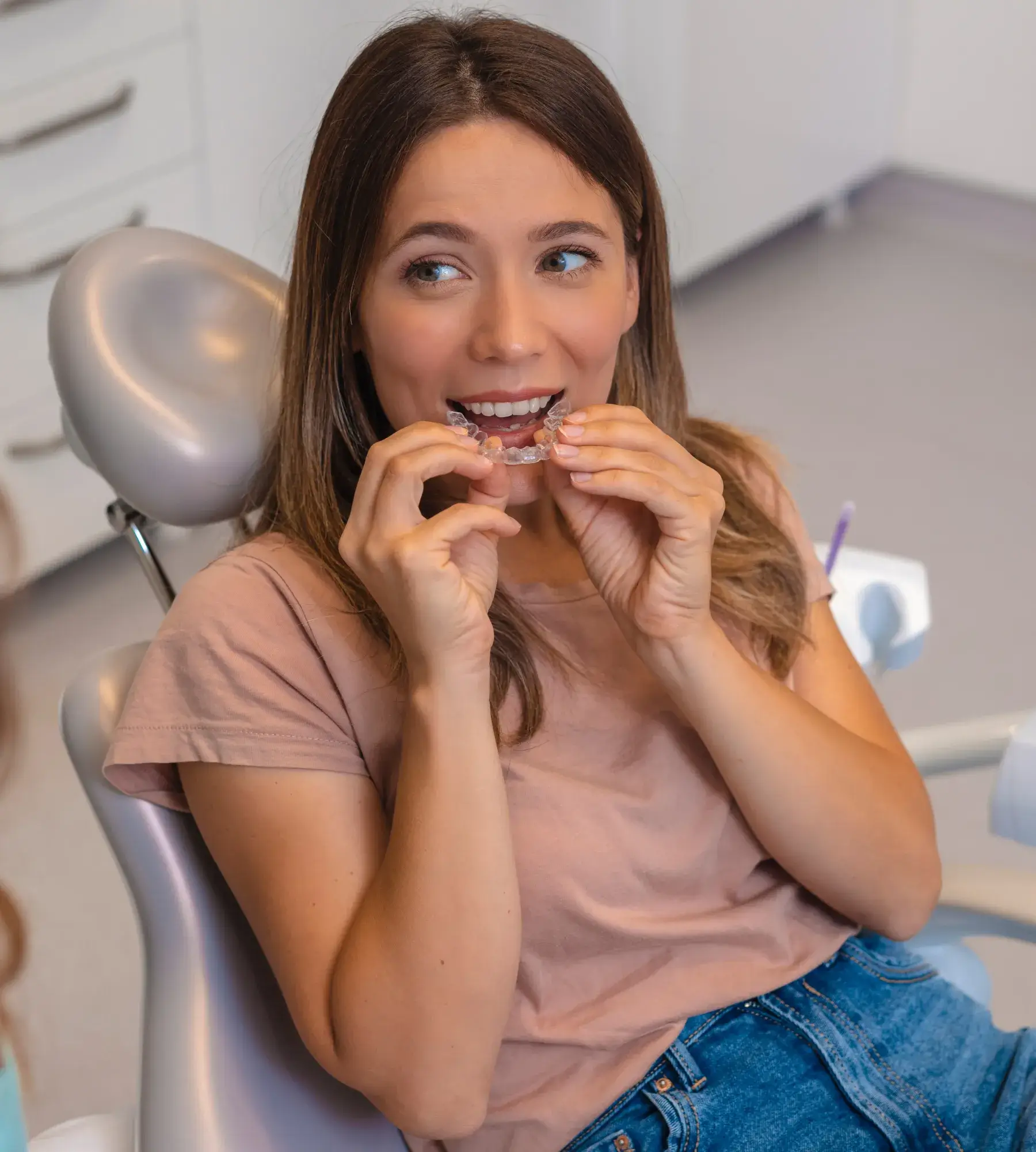 A woman sits in a dental chair, holding and looking at a clear aligner.
